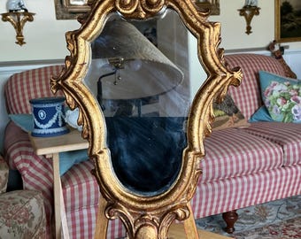 May include: A gold-colored, ornate mirror with an oval-shaped reflective surface. The mirror is supported by a small easel, and the frame features intricate scrollwork and decorative elements. The mirror is likely vintage or antique.