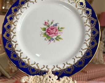 May include: A white porcelain plate with a blue and gold rim and a pink rose with green leaves in the center. The plate is on a white metal stand.
