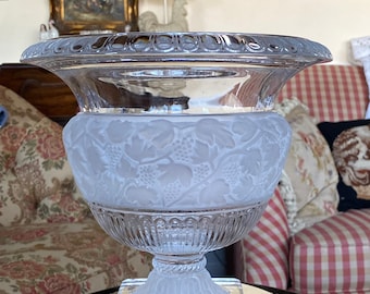 May include: A clear glass decorative bowl with a frosted grape design around the middle. The bowl has a wide, scalloped rim and sits on a square base. The bowl is displayed on a mirrored surface.
