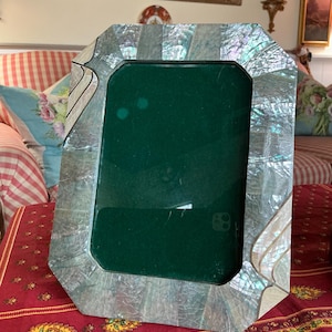 May include: An octagonal photo frame with a mother-of-pearl finish. The frame has a dark green backing and is set on a red patterned tablecloth. The frame is empty, ready to display a photograph.