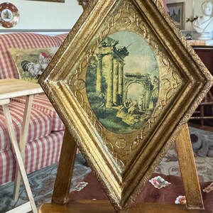 May include: A gold-framed picture with a scene of an ancient Roman ruin. The picture is on a wooden easel and is surrounded by a red and white checkered fabric.