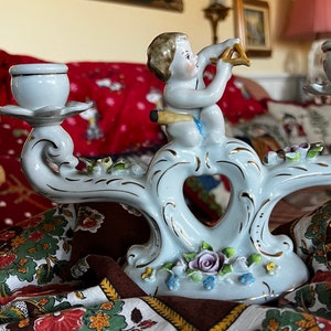 May include: A white porcelain candle holder with a heart-shaped design and a cherub figure playing a musical instrument. The holder has gold accents and floral details.