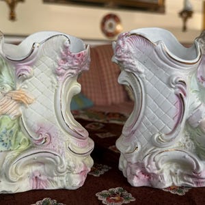 Puede incluir: Dos jarrones de porcelana con diseños florales ornamentados y figuras de mujeres en colores pastel. Los jarrones son blancos con detalles en rosa, verde y dorado.