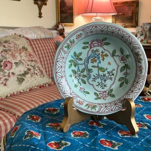 May include: A decorative, pale blue ceramic plate with a bird and floral design, displayed on a wooden stand. The plate features a detailed pattern with pink, green, and white accents. The plate is round and has a patterned border.
