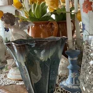 May include: A blue and green ceramic vase with a wavy rim and a textured surface. The vase is sitting on a wooden table with a crocheted doily.