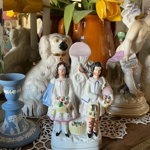 May include: A white porcelain figurine featuring two figures in dresses, each holding a basket of fruit. The figurine has a pink and blue color scheme, with gold accents. A white dog figurine and a blue candlestick are also visible.