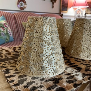 Puede incluir: Pantallas de lámpara beige con intrincados adornos de cuentas y lentejuelas. Las pantallas tienen forma cónica y están dispuestas sobre una mesa con un camino de mesa con estampado de leopardo. Decoración del hogar.