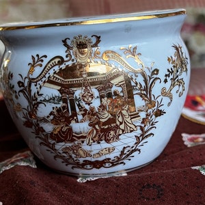 May include: A blue and gold ceramic planter featuring a decorative design of a scene with people in a building. The planter has a gold rim and a floral pattern.