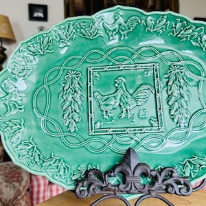 May include: A green ceramic serving dish with a raised design of roosters and leaves. The dish is oval shaped and has a scalloped edge.