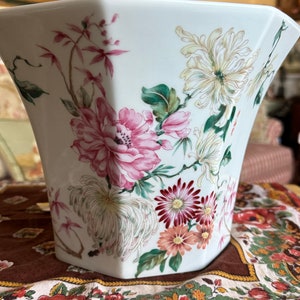 May include: White porcelain planter with a floral design featuring pink, white, and green flowers. The planter has a hexagonal shape and is sitting on a floral patterned fabric.