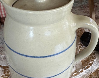 Puede incluir: Una jarra de cerámica blanca con tapa y diseño de rayas azules. La jarra tiene un asa y está sentada sobre un mantel de encaje blanco.