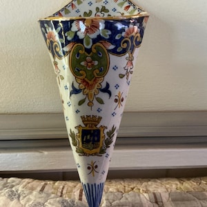 May include: A cone-shaped ceramic wall pocket vase featuring a white base with intricate hand-painted floral patterns in blue, green, and orange. The front displays a central heraldic crest with a crown, and the rim is finished with gold trim.