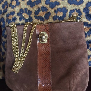 May include: A brown suede handbag with a gold chain strap and a decorative lion head clasp. The bag has a brown snakeskin panel on the front.