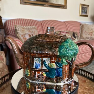 May include: A brown and green ceramic teapot shaped like a cottage with a thatched roof. The teapot has a scene painted on the side of two people looking out a window.