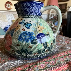 May include: A ceramic pitcher featuring a vibrant floral pattern with blue, green, and red blossoms, green leaves, and a blue dragonfly. The rim and handle are accented with a deep blue rose pattern, and the base has a decorative blue border.