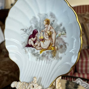 Puede incluir: Un plato blanco de porcelana con forma de concha y borde dorado, con una escena pintada de querubines. El plato está sostenido por un soporte ornamentado de color crema. Los querubines están representados con pelo y alas doradas.