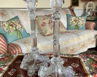 May include: A pair of clear glass candle holders with ornate designs. The candle holders have a tall, slender base with a wide, flared top. The base is decorated with intricate floral patterns and the top has a scalloped edge.