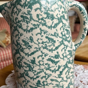 Puede incluir: Una jarra de cerámica verde y blanca moteada con asa. La jarra tiene un esmalte jaspeado y un aspecto rústico.