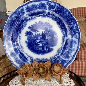 Puede incluir: Un plato de porcelana azul y blanco con un diseño floral y una escena de un castillo en un paisaje. El plato se muestra en un encaje de ganchillo blanco con un soporte floral de metal.