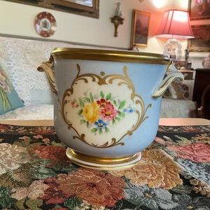 May include: A light blue porcelain cachepot featuring a central cream-colored cartouche decorated with a hand-painted floral bouquet of yellow, pink, and blue flowers. The vessel has gold-trimmed handles and a gold-painted rim and base.