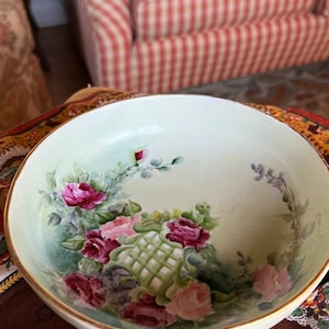 May include: A round, off-white ceramic bowl with a gold rim. The bowl is decorated with hand-painted pink and red roses, green leaves, and a lattice design. The bowl is sitting on a brown floral patterned tablecloth.