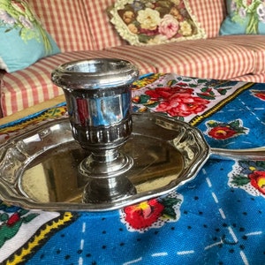 May include: Silver candle holder with a round base and a decorative handle. The holder is on a blue and white floral tablecloth.