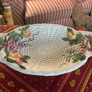 May include: White ceramic serving platter with woven texture and handles. The platter is decorated with colourful, raised relief fruit including grapes, peaches, and green leaves. A decorative piece for serving food.