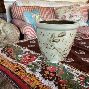 May include: A white ceramic planter with a brown leaf design and brown speckles. The planter is sitting on a brown and red floral tablecloth.