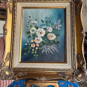 以下が含まれることがあります： 青い背景に白い花と紫の花のブーケが描かれた、額縁に入った油絵。フレームは金色で装飾が施されています。絵は小さなイーゼルの上に置かれています。