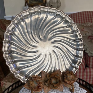 May include: A silver serving tray with a swirl design. The tray is sitting on a white crocheted doily with a decorative metal stand with three roses.