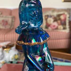 May include: A blue iridescent glass figurine of a woman with a gold trim around her neck and a white floral design on her dress.
