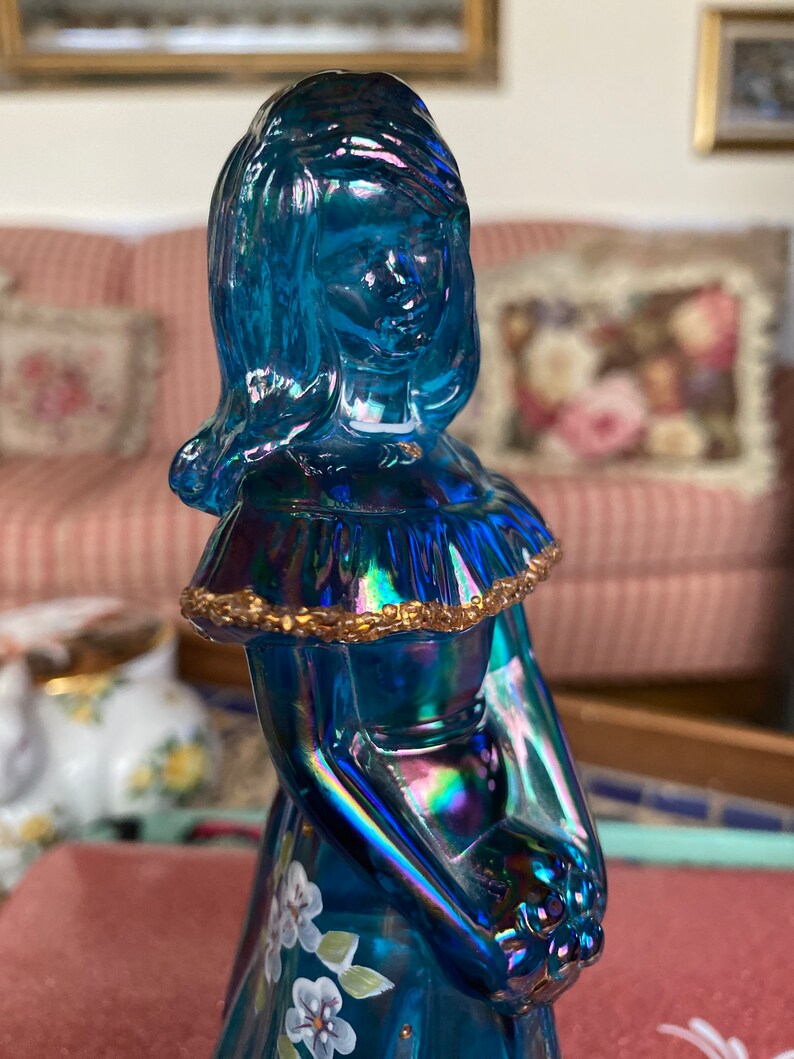 May include: A blue iridescent glass figurine of a woman with a gold trim around her neck and a white floral design on her dress.