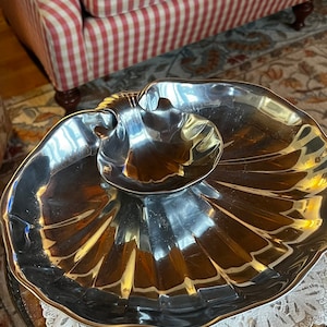 Puede incluir: Un plato de servir de plata con forma de concha marina con un tazón más pequeño en el centro. El plato está sobre una mesa redonda con un mantel de encaje blanco.