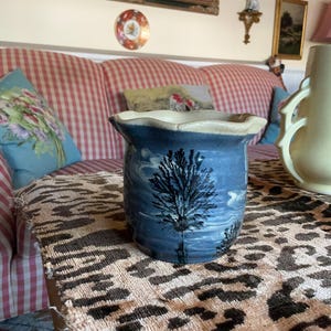 May include: A blue ceramic vase with a tree design. The vase has a wavy rim and a light-colored interior. The tree design is black and is set against a blue background. The vase is sitting on a table with a leopard print runner.