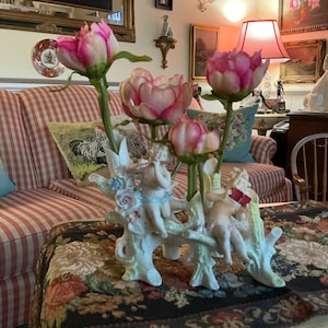 Könnte beinhalten: Dekorative Porzellan-Blumenarrangement mit rosa und weißen Blüten. Die Basis ist mit cherubischen Figuren verziert. Das Arrangement steht auf einem floral gemusterten Tisch, mit einem gestreiften Sofa im Hintergrund.