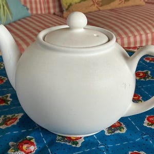 May include: A white ceramic teapot with a rounded body, curved spout, and a loop handle. The lid has a small knob and a vent hole. The teapot sits on a blue floral patterned tablecloth.
