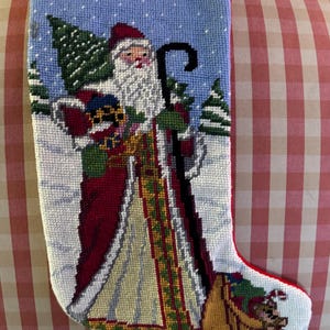 Puede incluir: Calcetín navideño de punto de cruz que representa a Papá Noel con una túnica roja y blanca, sosteniendo un bastón y regalos. El fondo muestra un paisaje nevado con árboles de hoja perenne y un cielo azul. El calcetín tiene un ribete rojo y un lazo para colgar.