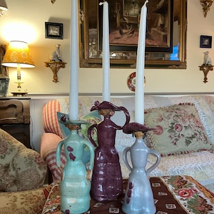 May include: Three ceramic candle holders shaped like women wearing hats. The candle holders are painted in shades of teal, purple, and gray. The candle holders are on a table with a floral tablecloth.