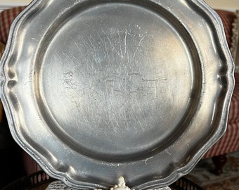 Puede incluir: Un plato de peltre plateado con un borde festoneado, que muestra signos de desgaste y uso. El plato se muestra en un encaje de ganchillo blanco.
