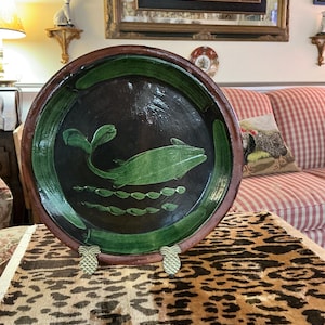 May include: A large, round, ceramic plate with a dark brown rim and a black interior. A green fish design is painted in the center, with green accents around the edge. The plate is displayed on a leopard print surface.