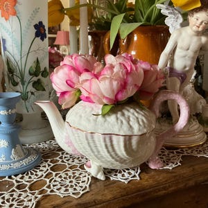 May include: A white porcelain teapot with a pink and white floral design. The teapot has a shell-like design and is sitting on a white doily. The teapot is surrounded by other decorative items, including a porcelain figurine of a cherub.