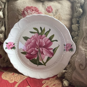 Könnte beinhalten: Weiße Keramik-Servierplatte mit einem Design aus rosa Pfingstrosen. Die Platte hat einen gewellten Rand und zwei Griffe.