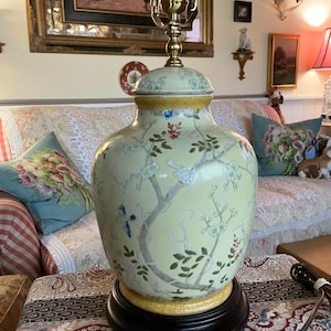 Peut inclure: Une lampe de table décorative avec une base et un couvercle en céramique jaune pâle, ornée d'un motif floral et d'oiseaux peint. La lampe repose sur une base en bois noir et possède un support de lampe en laiton.