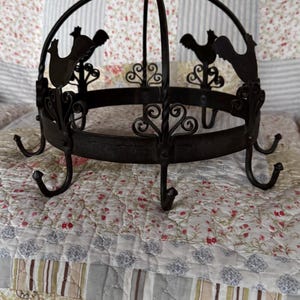 May include: A dark brown metal pot rack featuring a circular frame with multiple downward-facing hooks. The structure is adorned with decorative scrollwork and silhouettes of roosters positioned around the upper rim, topped with a central hanging loop.