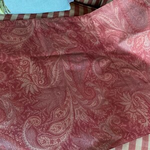 May include: A section of red fabric with a paisley pattern in a lighter shade of red. The fabric is folded, showing the intricate design of the paisley. The fabric is likely used for clothing or home decor.
