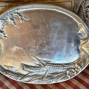Puede incluir: Una bandeja de servir de plata con un diseño ornamentado con mazorcas de maíz y flores. La bandeja tiene dos asas en forma de concha.