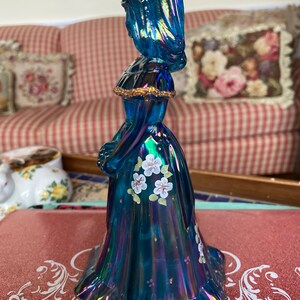 May include: A blue iridescent glass figurine of a woman in a long dress with white flowers painted on the skirt.
