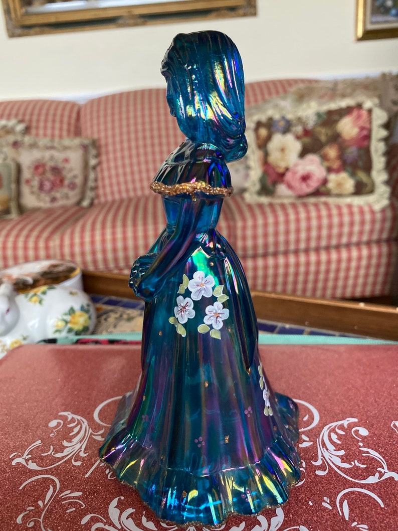May include: A blue iridescent glass figurine of a woman in a long dress with white flowers painted on the skirt.