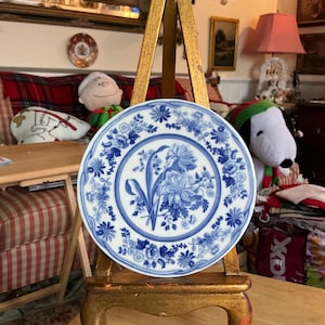 May include: A white and blue decorative plate with a floral design. The plate features a central bouquet of flowers and is surrounded by a border of smaller floral patterns. The plate is displayed on a gold-colored easel.