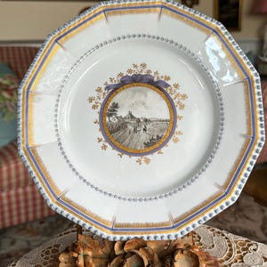 May include: A white porcelain plate with a blue and gold rim and a central design of a country scene with a house and figures. The plate is on a decorative stand with floral accents.